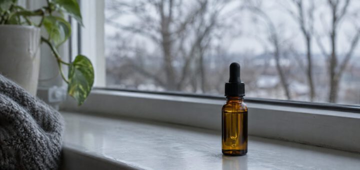 Vitamin-D-Tropfen in einer braunen Pipettenflasche auf der Fensterbank, draußen kahle Bäume im Winterlicht