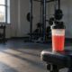Shaker mit rotem Pre-Workout-Booster auf einer Hantelbank im Studio, dahinter Kurzhantelablage und Power Rack
