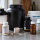 Supplements im Test: Proteindose, Shaker mit Eiweißshake, Messlöffel und mehrere Kapseldosen auf der Küchenarbeitsplatte