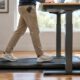 Füße in Laufschuhen auf einem flachen Walking Pad unter dem Steh-Schreibtisch, das Handy steckt in der Hosentasche